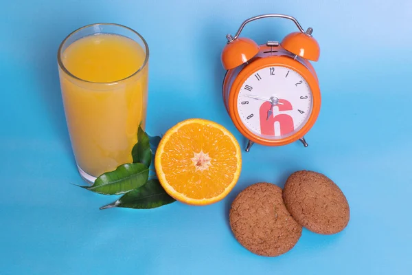 Closeup of an orange alarm clock with a glass of orange juice, fresh ripe fruit cut in half, oatmeal cookies on a blue background. Organic and healthy breakfast concept.
