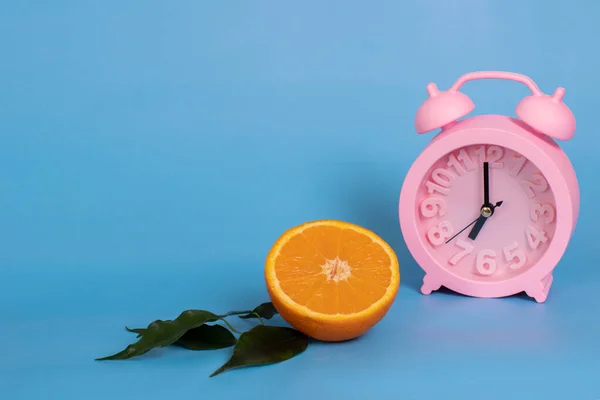 Pinkfarbener Wecker und ein halbes Orange auf blauem Hintergrund. Das Konzept der Sommergetränke, gesunde Ernährung. Kopierraum — Stockfoto