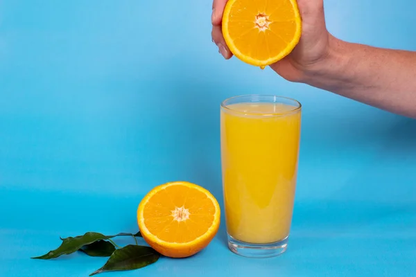 Hand squeezing juice from half an orange, orange juice in a glass, half an orange with leaves on blue background.