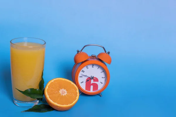 Fresh orange juice in a glass, fruit cut in half and sliced with green leaves, orange alarm clock isolated on a blue background, copy space. Time to drink juice