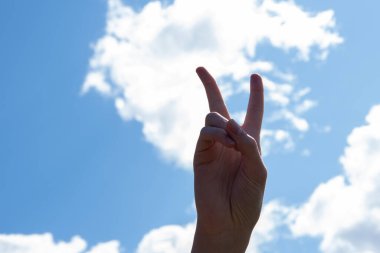 Barış ya da zaferin sembolü olarak iki parmağını kaldırarak, işaret dilinde V harfinin işareti. Mavi gökyüzü arka planında bulutlarla izole edilmiş, boşluğu kopyala.