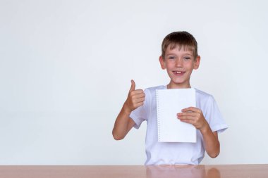 Beyaz bir not defteri tutan beyaz bir beyaz beyaz çocuğun portresi, baş parmaklarını gösteriyor. Beyaz arka planda, ışık masasında oturan bir çocuk. Okula dönüş kavramı, neşe, mutluluk. Boşluğu kopyala