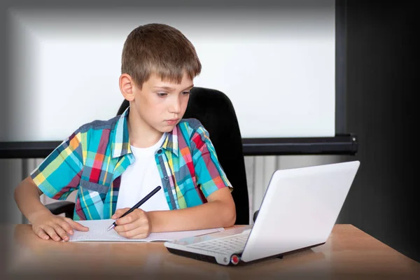Un chico lindo está sentado en una mesa, mirando un portátil, escribiendo tareas o preparándose para un examen. Concepto Volver a la escuela — Foto de Stock