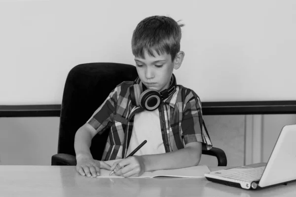 Un chico lindo está sentado en una mesa, mirando un portátil, escribiendo tareas o preparándose para un examen. Concepto Volver a la escuela — Foto de Stock