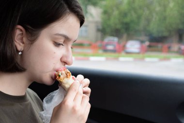 Genç ve güzel bir kadın arabada fast food yiyor. Ayrıl. Akşam yemeği arabada. Ön koltukta. Yemek konsepti. Sağlıksız gıda konsepti, araba kullanma, fast food.
