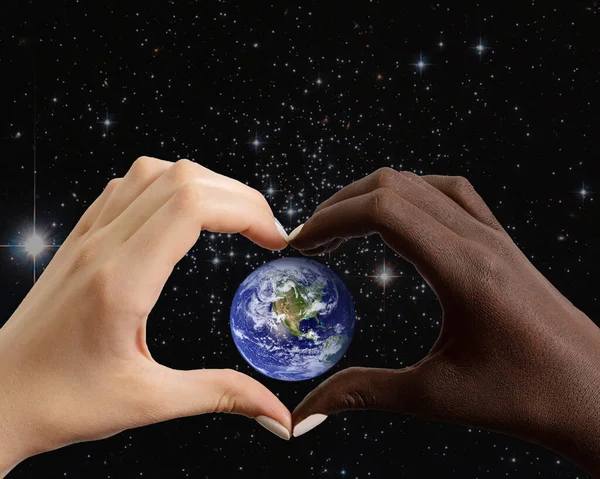 Manos blancas y negras en forma de corazón con el planeta Tierra sobre el fondo del cielo estrellado —  Fotos de Stock