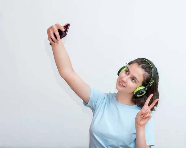 Chica bastante caucásica en una camiseta azul y auriculares hace una selfie utilizando una aplicación en un teléfono inteligente sobre un fondo blanco — Foto de Stock