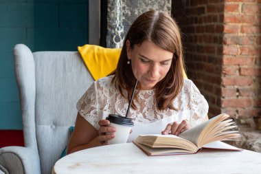 Beyaz bluzlu, kızıl uzun saçlı güzel bir kadın kafede bir sandalyede tek başına oturur, kamışla kahve içer ve kitap okur.