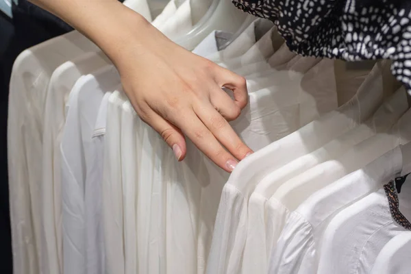 girl chooses a white shirt in the store. Selection of new clothes close-up of hands. Clothes on hangers in a store for sale. Selection of clothes in the store. Shopping concept, business concept