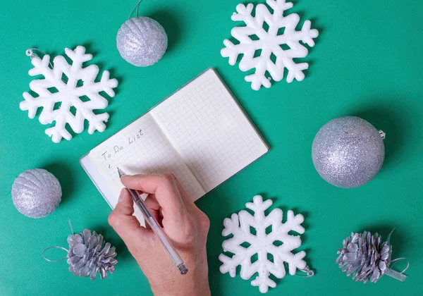 A female hand writes a to-do list in a blank notebook that needs to be done before Christmas, silver balls, white snowflakes, pine cones on a green background