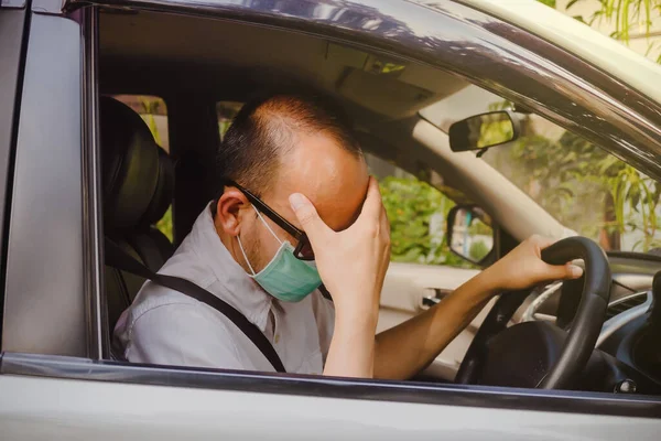 Coronavirus and Air pollution pm2.5 concept. Man driving wearing protective mask to avoid the infection of virus and polluted air spreading deseases, holding his head, having a headache, driving a car