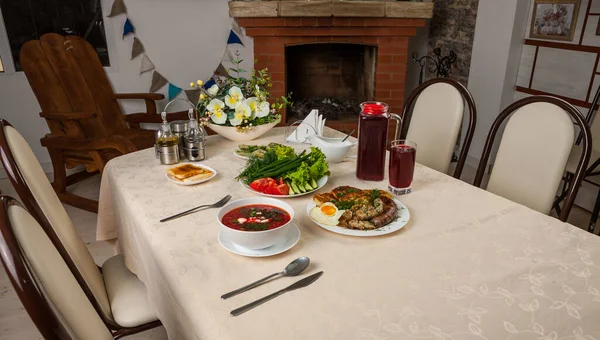 Russian lunch with red borsch, fresh vegetables, fried egg, fried sausage, bacon and potato pancakes. Closeup photo of Russian hot main meal. Loft interior of dining room. Brick fireplace, rough wooden arm-chair.