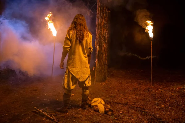 The man with long hair in textile long shirt is looking to the forest. Viking's nighttime in the forest is lighting by flamed torches.
