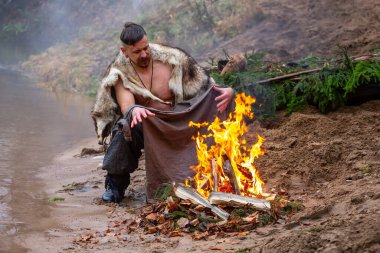 Zaslavl, Belarus, 10.11.2019. Ukraynalı adam rafting yaptıktan sonra gömleğini ateşin yanında kurutur. Zaporozhian Kozak kendini kızgın ateş ve kürk derisi ile ısıtıyor. Manzara, sonbahar zamanı.