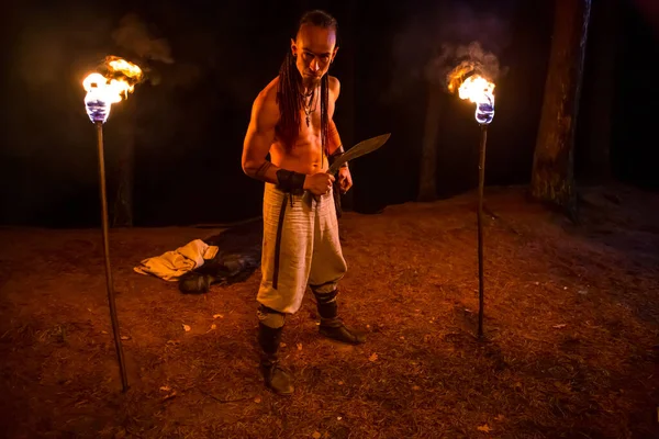 Irdorath group, clip making backstage. The caucasian man with dreadlocks,  with knife in the hand. Viking\'s nighttime in the forest is lighting by flamed torches.