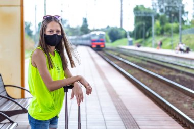 İyi yolculuklar. Siyah maskeli, bavullu beyaz bir genç tren istasyonunda trenini bekliyor.. 