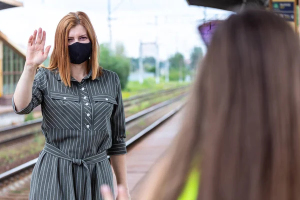 Caucasian female in a black face mask say hello by the hand at the railway station. Safe travel.