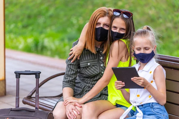 Safe family travel. Mother and two daughters in face masks are waiting for take off by train at the railway station. City view.