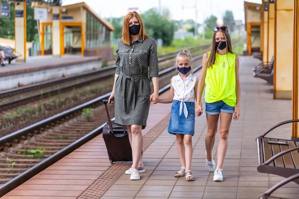 Safe family travel. Mother and two daughters in face masks are waiting for take off by train at the railway station. City view.