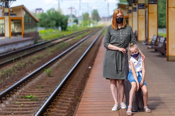 Caucasian female with daughter in a black face masks with luggage are waiting for take off at the railway station. Safe travel.