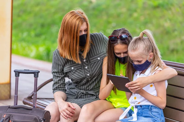 Safe family travel. Mother and two daughters in face masks are waiting for take off by train at the railway station. City view.