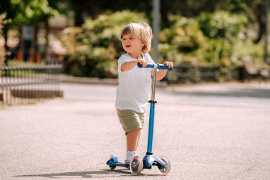 Çocuk güneşli bir yaz gününde parkta scooter sürmeyi öğreniyor. Anaokulu çocuğu paten kayıyor. Çocuklar dışarıda scooterlarla oynarlar. Çocuk için aktif eğlence ve açık hava sporu.