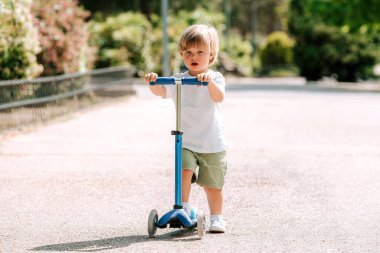 Çocuk güneşli bir yaz gününde parkta scooter sürmeyi öğreniyor. Anaokulu çocuğu paten kayıyor. Çocuklar dışarıda scooterlarla oynarlar. Çocuk için aktif eğlence ve açık hava sporu.