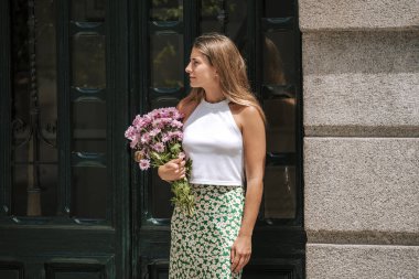 Çiçek buketi olan güzel bir kadın evin kapısında bekliyor. Sokakta portresi olan güzel bir genç kız..