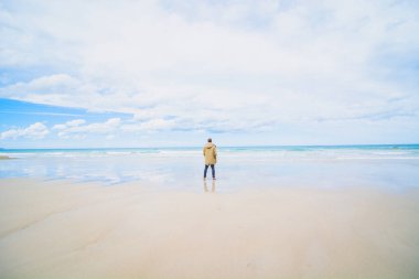 Kolları açık genç bir adam, çoğunlukla bulutlu bir günde okyanusu düşünüyor.