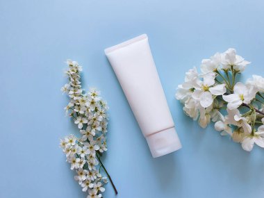 Yüz nemlendirici kremi ya da açık mavi arka planda yüz temizleyicisi olan beyaz plastik bir tüp. Çiçek süsleme, düz döşeme ve Üst Manzara.