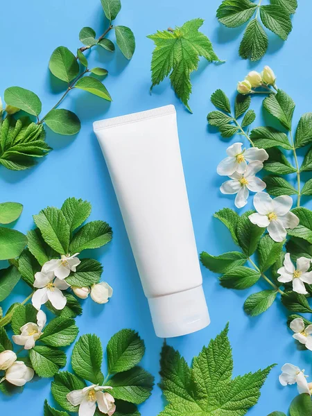 Cosmetics SPA branding mock-up with fresh green leaves and white flowers on a deep blue background. White plastic tube for branding, flower flat lay. Vertical photo.