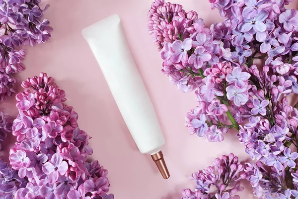 Top view of face and eye cream squeeze, cosmetic tube with long nozzle and bronze screw cap and pink lilac flowers on a powdery pink background. Cosmetics spa branding mockup, flower flat lay.