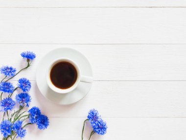 Beyaz bir fincan kahve ve beyaz ahşap arka planda mavi çiçek. Güzel bir yaz yatağı. Metnin ve ürünün için alanı kopyala.