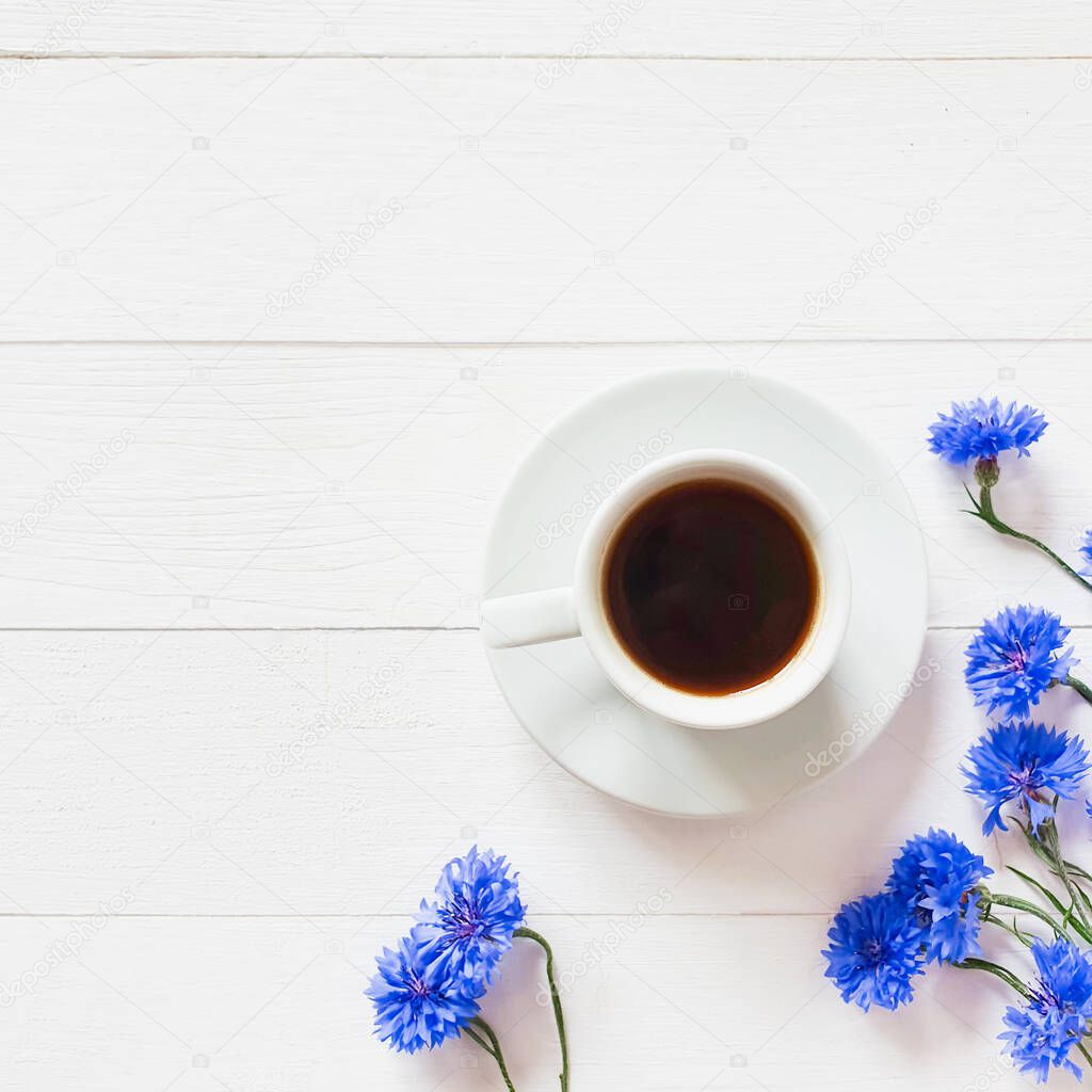 White cup of coffee and blue cornflowers on a vintage white wooden background. Lovely summer flat lay. Copy space for your text and product. Top view, square photo.