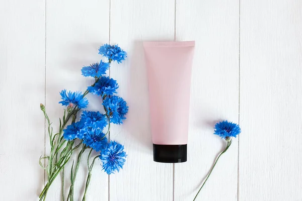 Top view of mockup pink squeeze bottle plastic tube with black cap and blue cornflowers on a vintage white wooden background. Natural organic spa cosmetics concept. Lovely summer flat lay.