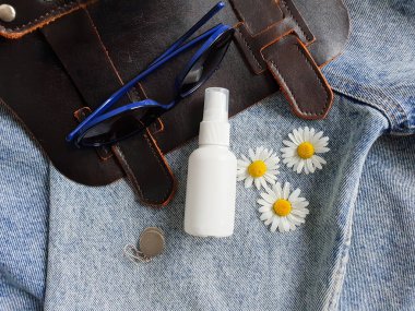 Kotun arka planında markasız beyaz plastik sprey sprey şişesi ve el çantası, güneş gözlüğü, papatya ve gümüş küpelerin en üst görüntüsü. Damgalamak için şişe.