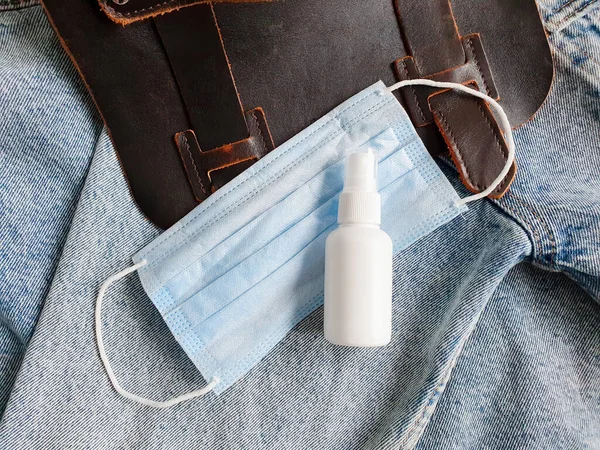 Bottle of hand sanitizer, antimicrobial liquid spray, medical protective mask and brown bag on blue denim background. Top view, mock-up. Bottle for branding.