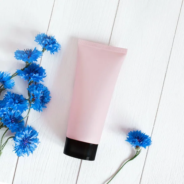 Top view of mock-up pink squeeze bottle plastic tube with black cap and blue cornflowers on a white wooden background. Natural organic spa cosmetics concept. Flatlay, square photography.