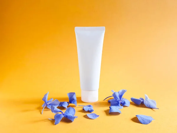 Mockup of unbranded white bottle for branding and label and petals of blue hydrangea flowers on a textured bright orange yellow background. Squeeze bottle plastic tube. Natural organic spa cosmetics