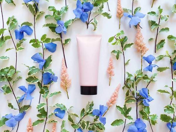 Top view of mockup of pink squeeze bottle plastic tube with black cap, fresh greens, blue and pink flowers on a white background. Bottle for branding and label. Natural organic spa cosmetics concept.