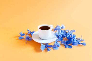 Bir fincan kahve ve mavi ortanca çiçekleri dokulu sarı-turuncu arka plan.