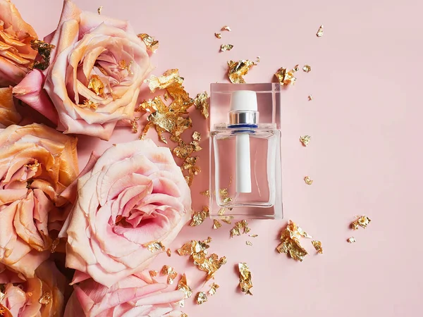 Perfume bottle, pink gold roses and pieces of golden paper on powdery pink background. Top view, flower flatlay.