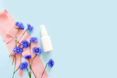 Açık mavi arka planda beyaz plastik sprey ve mavi çiçeklerin maketi. Doğal organik spa kozmetik ürünleri, sıvı antimikrobik sprey konsepti. Çiçek düzlüğü, üst görünüm, kare fotoğraf, kopyalama alanı.