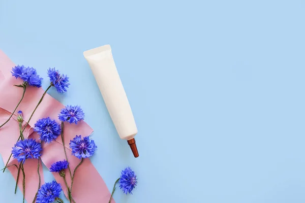 Mockup of ivory cosmetic tube with long nozzle and bronze screw cap, silk ribbons and blue flowers on a pastel blue background. Face and eye cream squeeze bottle. Natural organic spa cosmetics