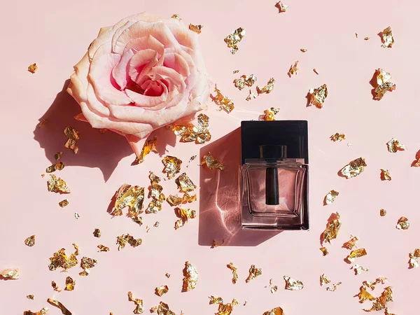 Siyah şapkalı dikdörtgen cam parfüm şişesi, pembe altın gül ve pastel pembe masa üzerinde altın kağıt parçaları. Moda gölgesi olan lüks düz yatak.