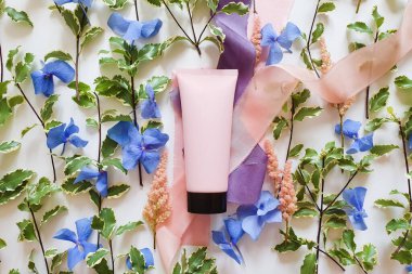 Siyah kapaklı pembe plastik tüp, taze yeşiller, mavi ve pembe çiçekler, beyaz arka planda ipek kurdeleler. Damgalamak ve etiketlemek için bir şişe. Doğal organik kaplıca konsepti.