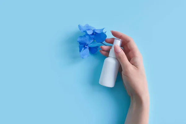 Mock-up of white plastic spray bottle, female hand and blue flowers on a blue background. Natural organic spa cosmetics and liquid antimicrobial spray concept.