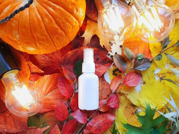 Mockup of white plastic spray bottle pumpkin, magic lights and bright autumn leaves. Atmospheric fall flatlay. Natural organic spa cosmetics and liquid antimicrobial spray concept. Mock-up, top view.
