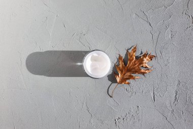 Açık cam kavanozda nemlendirici krem gri arka planda altın yaprak. Moda uzun gölgesi, üst görünüm, kopyalama alanı.