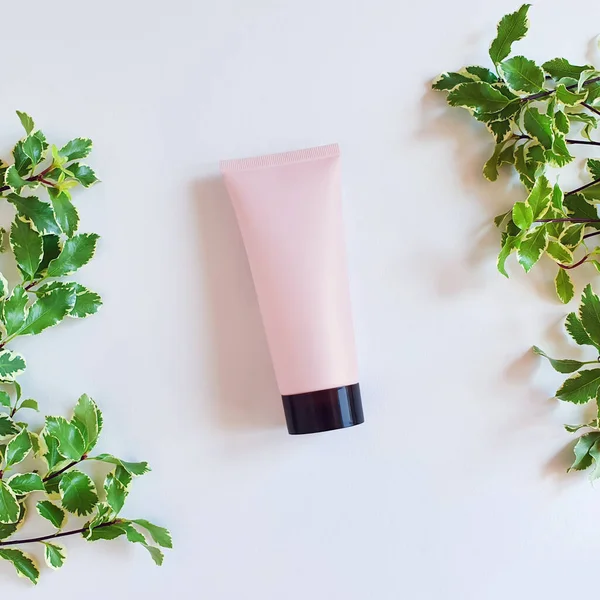 Mockup of pink squeezed cosmetic tube with black cap and fresh greens on white table. Bottle for branding and label. Natural organic spa cosmetics concept. Top view, flat lay style.